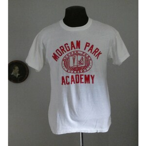 May include: White t-shirt with a red print of the Morgan Park Academy logo. The logo is a circle with the words "Morgan Park Academy" around the outside and a book, torch, and compass inside.