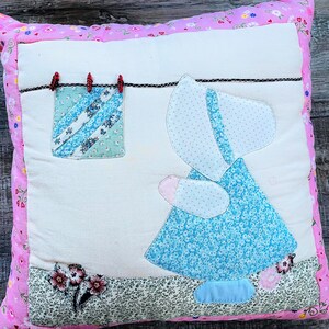 May include: Square pillow with a patchwork design featuring a girl in a blue dress and bonnet. A small square of fabric hangs on a clothesline. The pillow has a pink floral border and a white center.