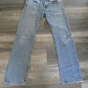 May include: Light blue denim jeans with a distressed look, featuring a button fly and a ripped knee. The jeans are laid flat on a wooden surface, showcasing their classic five-pocket design and faded areas.