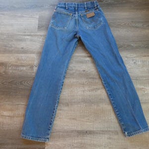 May include: Blue denim jeans with a classic five-pocket design. The jeans have a straight leg cut and a brown leather patch on the back pocket. The jeans are laid flat on a wooden surface.