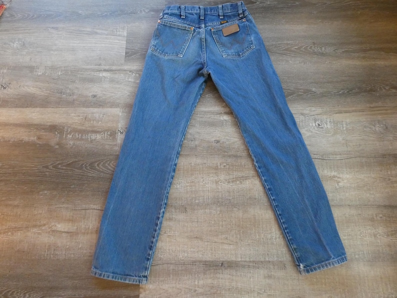 May include: Blue denim jeans with a classic five-pocket design. The jeans have a straight leg cut and a brown leather patch on the back pocket. The jeans are laid flat on a wooden surface.