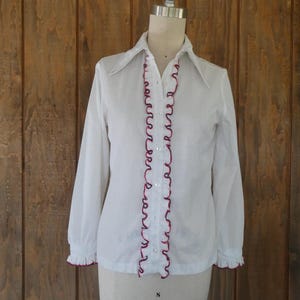 May include: White long-sleeve blouse with a pointed collar and button-down front. The blouse features ruffled trim in a deep red color along the placket and cuffs. The garment is displayed on a mannequin against a wooden background.