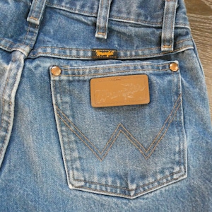 May include: Close-up of blue denim jeans with a Wrangler label on the back pocket. The jeans have copper-colored rivets and a brown leather patch. The stitching is in a contrasting tan color.