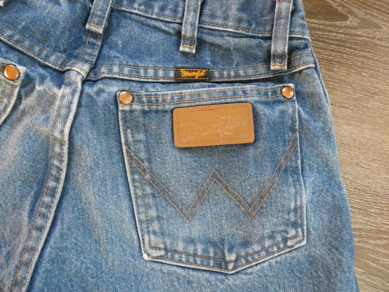 May include: Close-up of blue denim jeans with a Wrangler label on the back pocket. The jeans have copper-colored rivets and a brown leather patch. The stitching is in a contrasting tan color.