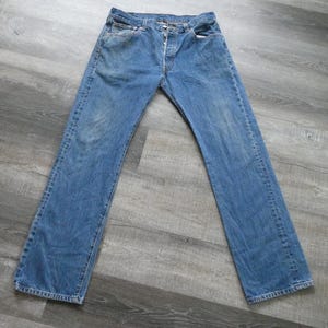 May include: Light blue denim jeans with a button fly. The jeans have a classic five-pocket design and a straight leg cut. The denim has a faded appearance, suggesting wear and washing. The jeans are laid flat on a gray surface.