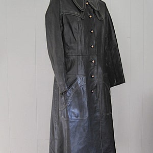 May include: A long black leather coat with a white stitched trim around the collar and pockets. The coat has a button closure and a fitted silhouette.