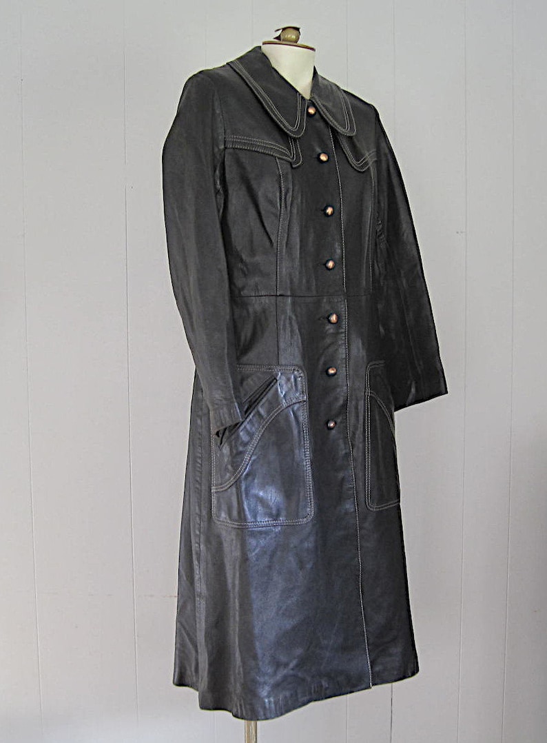 May include: A long black leather coat with a white stitched trim around the collar and pockets. The coat has a button closure and a fitted silhouette.