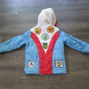 May include: A vintage children's winter jacket with a white hood, light blue sleeves, and a red center panel. The jacket features various colorful patches, including a Delta logo and cartoon character designs. The jacket is laid flat on a wooden surface.