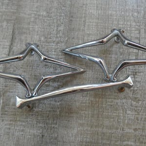 May include: Three silver-toned metal drawer pulls with a distinct diamond-shaped, open-frame design. Each handle features a polished, reflective surface and pointed ends, with visible mounting points at the base for attachment to furniture.