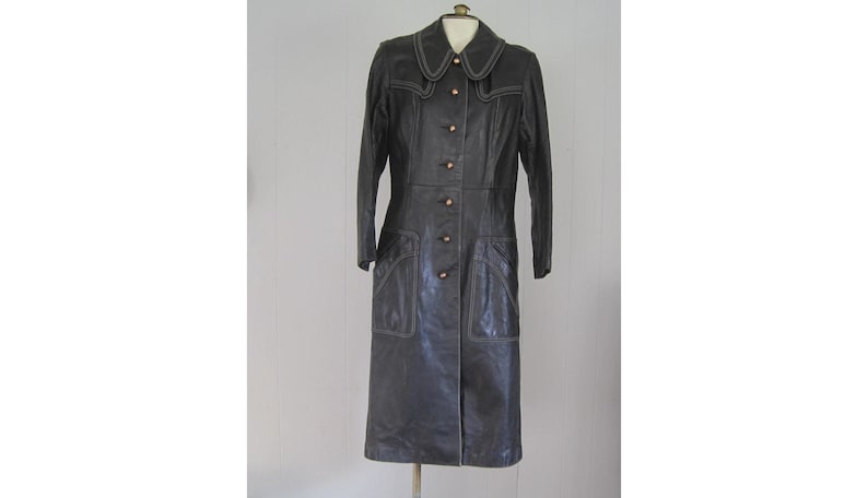 May include: A long black leather coat with a white stitching detail and a button closure. The coat has a collar and two pockets.