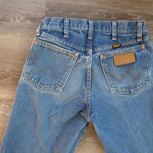 May include: Close-up of the back of blue denim jeans. The jeans have two back pockets with a signature stitching design and a brown leather patch with the Wrangler logo. The jeans are a light blue wash.