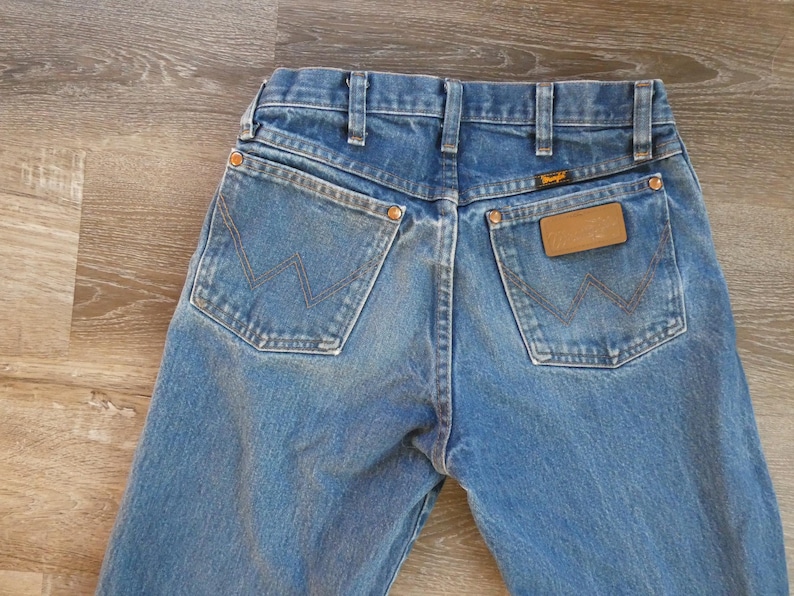 May include: Close-up of the back of blue denim jeans. The jeans have two back pockets with a signature stitching design and a brown leather patch with the Wrangler logo. The jeans are a light blue wash.