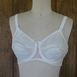 May include: White vintage bra with floral lace detailing. The bra features adjustable straps and a delicate lace trim along the neckline and underbust. The bra is displayed on a mannequin against a wooden background.