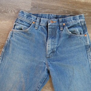 May include: Close-up of blue denim jeans. The jeans have a classic five-pocket design with copper rivets and a button closure. The denim has a faded wash, giving it a vintage look. The jeans are laid flat on a wooden surface.