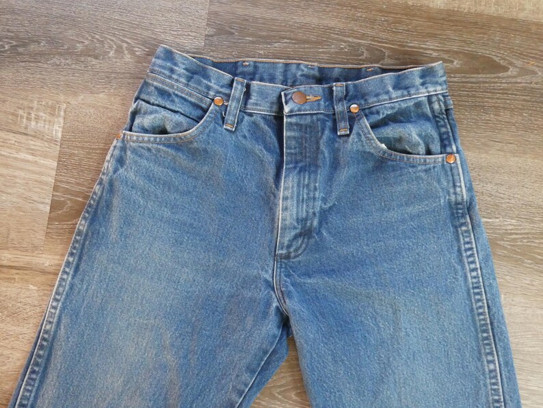 May include: Close-up of blue denim jeans. The jeans have a classic five-pocket design with copper rivets and a button closure. The denim has a faded wash, giving it a vintage look. The jeans are laid flat on a wooden surface.