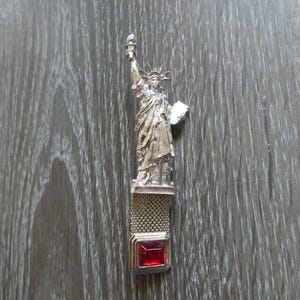 May include: Silver-toned Statue of Liberty charm with a red rhinestone. The charm is attached to a silver chain.