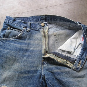 May include: A pair of blue denim jeans with a zip fly and a button fastening. The jeans have a faded and distressed look, with visible wear and tear. The jeans are open at the fly, revealing the white lining.