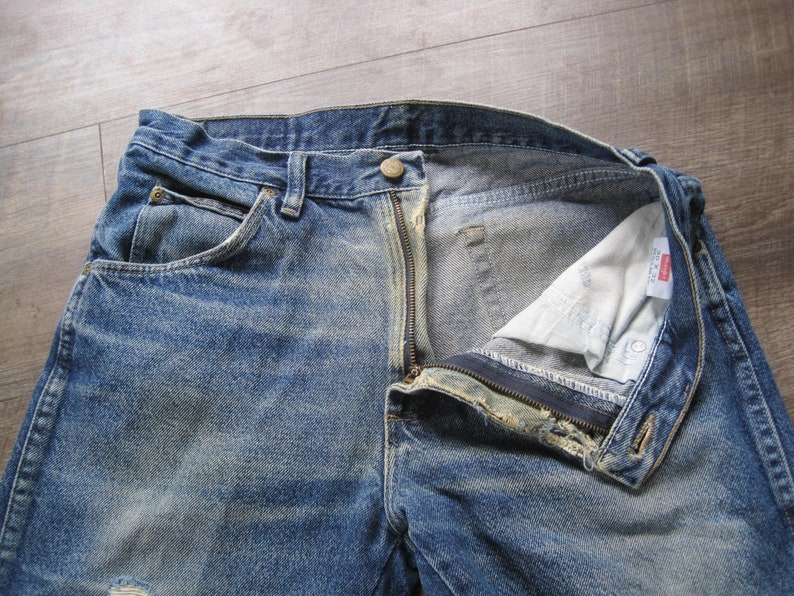 May include: A pair of blue denim jeans with a zip fly and a button fastening. The jeans have a faded and distressed look, with visible wear and tear. The jeans are open at the fly, revealing the white lining.