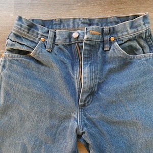 May include: Close-up of blue denim jeans with a zipper fly, button closure, and front pockets. The jeans have a classic, worn look, with visible stitching and metal rivets.