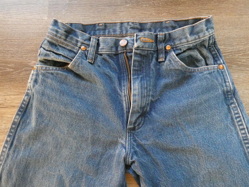 May include: Close-up of blue denim jeans with a zipper fly, button closure, and front pockets. The jeans have a classic, worn look, with visible stitching and metal rivets.