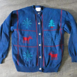 May include: A blue knitted cardigan with a Christmas theme. The cardigan has a button-up front and features red, green, and white snowflakes, reindeer, and Christmas trees.