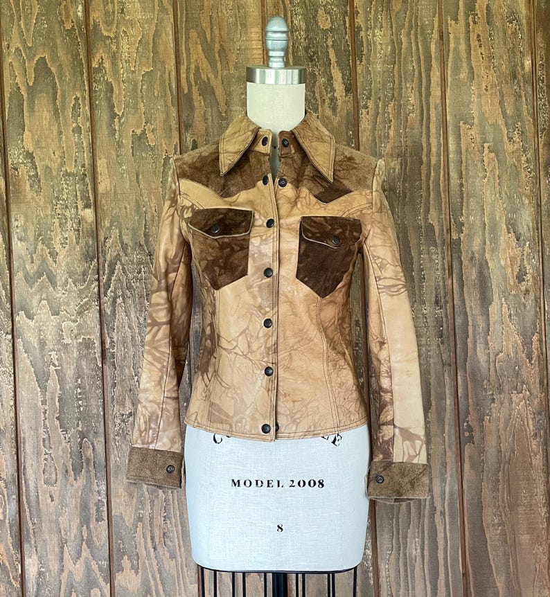 Peut inclure: Une veste en cuir marron avec des accents en daim. La veste a une fermeture boutonn&eacute;e &agrave; l'avant et des manches longues. La veste est sur un mannequin blanc.