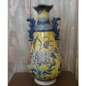 May include: A tall, yellow ceramic vase with a floral design and two blue handles. The vase features a large gray and white peony, with other flowers and leaves. The top and handles are dark blue, and the base is white.