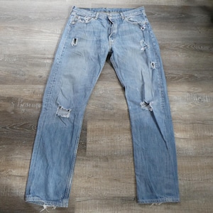 May include: Light blue denim jeans with distressed details, including rips at the knees and thighs. The jeans have a classic five-pocket design and a button-and-zipper closure. The jeans are laid flat on a wooden surface.