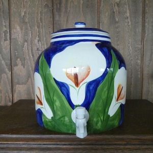 May include: A ceramic beverage dispenser with a lid, featuring a hand-painted floral design. The white flowers with orange centers are set against a blue background with green leaves. The dispenser has a white spigot and is on a wooden surface.