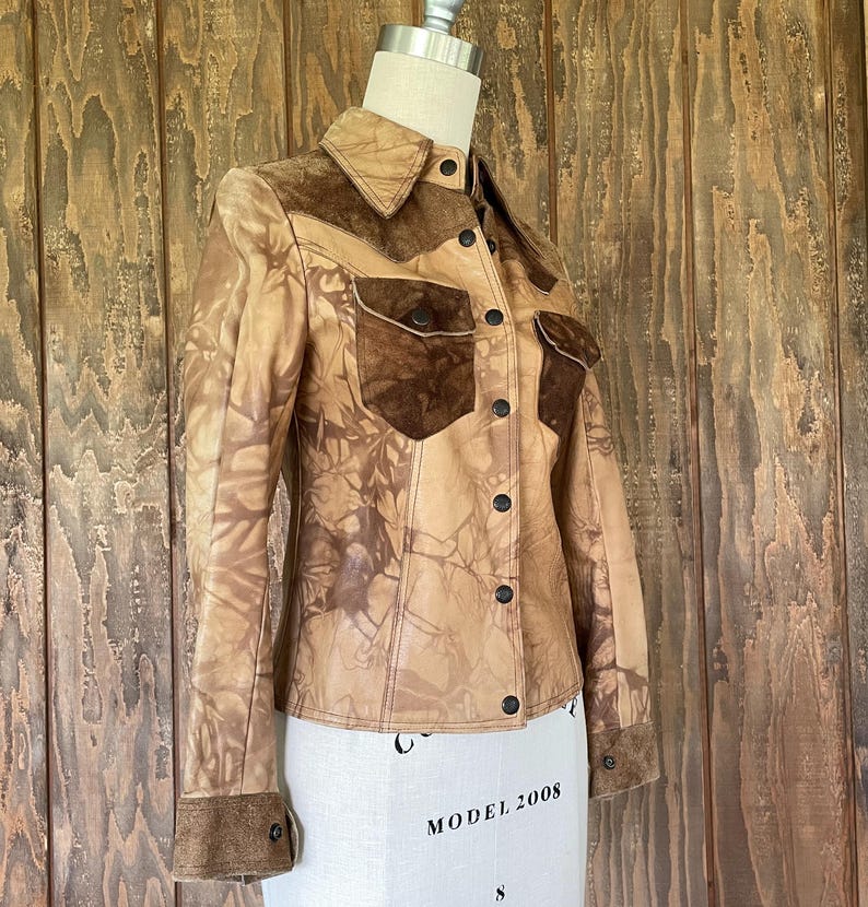 Peut inclure: Une veste en cuir marron avec un col et des poches en daim. La veste a un motif tie-dye et une fermeture boutonn&eacute;e.