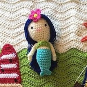 Puede incluir: Una mu&ntilde;eca de sirena de crochet con cabello azul, cola verde y una flor rosa en la cabeza. La mu&ntilde;eca est&aacute; sentada sobre una superficie de crochet verde.