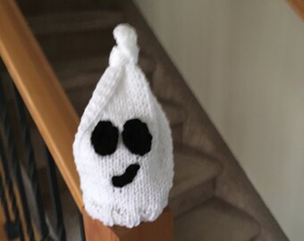 Puede incluir: Sombrero de fantasma de punto blanco con ojos de fieltro negro y una boca sonriente. El sombrero está sentado en una barandilla de madera.