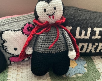 May include: A handmade crochet vampire doll featuring a white face with black eyes and red fangs, black hair, a grey shirt, black pants, and a bright red cape tied around the neck. The doll has white hands and is crafted with visible yarn stitches.