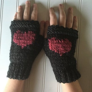 May include: A pair of black fingerless gloves with a red heart design knitted into the center. The gloves are made of a textured yarn and have a ribbed cuff. The background is a white paneled wall.