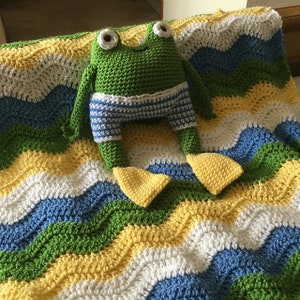 May include: A crocheted green frog with white and blue striped shorts sits on a crocheted blanket with yellow, white, blue and green wavy stripes.