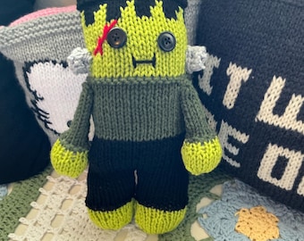 May include: A handmade knitted doll resembling a green monster with black hair, black button eyes, and a red stitched scar on its forehead. The doll features light gray bolts on the sides of its head, a dark green shirt, and black pants with lime green feet.