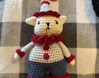 May include: A crocheted stuffed animal toy featuring a cream-colored head and limbs, a white torso, and blue pants. The toy wears a red ruffled collar, red wrist cuffs, red ankle cuffs, and a pointed cream hat with a red brim and red pom-pom.