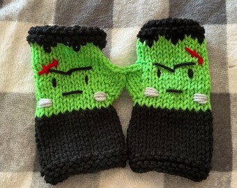 May include: A pair of fingerless gloves knitted with bright green yarn featuring a character face with black hair, black eyebrows, a straight mouth, and two grey stitches. Each glove has a black cuff and a small red stitched scar on the forehead.
