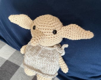 May include: A beige crochet doll with large pointed ears, black bead eyes, and a small snout. The doll is wearing a light gray knitted tunic with a simple tie detail at the shoulder, crafted with visible yarn stitches and a soft, rounded head shape.