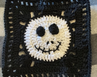 May include: A square crochet granny square featuring a circular white face in the center with two black circular eyes and a stitched black mouth. The surrounding border is made of black yarn with an open, airy, geometric grid pattern.