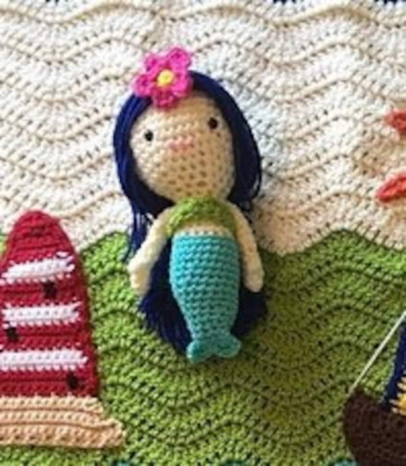 Puede incluir: Una mu&ntilde;eca de sirena de crochet con cabello azul, cola verde y una flor rosa en la cabeza. La mu&ntilde;eca est&aacute; sentada sobre una superficie de crochet verde.
