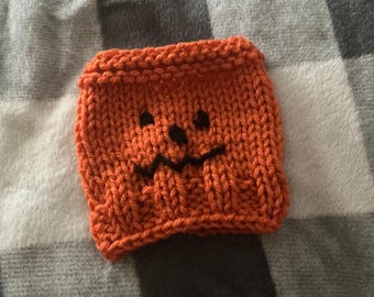 May include: A square, orange knitted cozy featuring a hand-stitched black yarn face with two circular eyes, a small nose, and a jagged, zig-zag mouth. The item is constructed with a ribbed knit texture and finished with a rolled top and bottom edge.