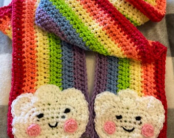 Può includere: Una sciarpa all'uncinetto arcobaleno con due nuvole bianche all'uncinetto attaccate alle estremità. Ogni nuvola ha un viso sorridente con guance rosa.