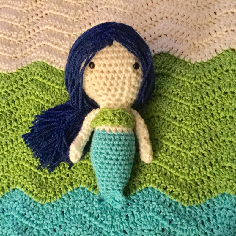 Puede incluir: Una mu&ntilde;eca de sirena de crochet con cabello azul, cara blanca y cola verde y azul. La mu&ntilde;eca est&aacute; acostada sobre una manta de crochet verde y azul.