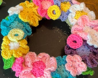 May include: A circular wreath decorated with numerous crocheted flowers in vibrant shades of pink, yellow, blue, purple, and white. The floral arrangement features various petal shapes and sizes, interspersed with small green crocheted leaves.