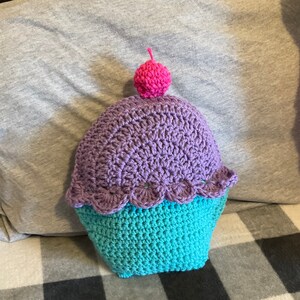 Può includere: Un cuscino a forma di cupcake all'uncinetto con glassa viola e una ciliegia rosa in cima. Il cuscino è turchese in basso e ha un bordo smerlato.
