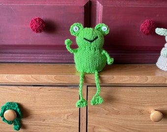 Op de afbeelding: Een groene gebreide kikker met witte ogen en een glimlachende mond zit op een houten dressoir met twee lades. Het dressoir heeft een groene klaverbladvormige knop op de bovenste lade en een ronde knop op de onderste lade.