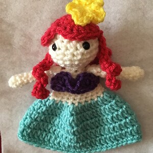 Puede incluir: Una mu&ntilde;eca de ganchillo de una sirena con cabello rojo, un top morado y una cola turquesa. La mu&ntilde;eca lleva una corona de flores amarilla.