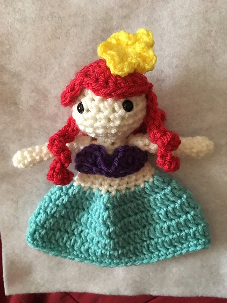 Puede incluir: Una mu&ntilde;eca de ganchillo de una sirena con cabello rojo, un top morado y una cola turquesa. La mu&ntilde;eca lleva una corona de flores amarilla.