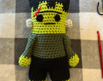 May include: A handmade crochet doll featuring a lime green head with black hair, black eyes, and a red stitched scar. The body is dark olive green with black pants and lime green hands and feet, accented by small light gray side bolts.